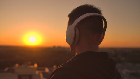 Close-up-of-a-man-in-headphones-looking-at-the-city-from-the-height-of-a-skyscraper-at-sunset.-Relax-while-listening-to-music.-Enjoy-a-beautiful-view-of-the-city-at-sunset-from-the-roof-with-headphones.